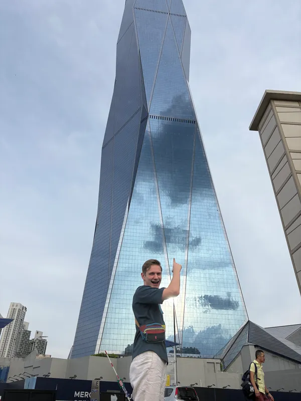 Mathew grinning and pointing up at Merdeka 118, a twisting glass skyscraper in Kuala Lumpur
